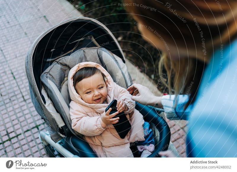 Bezauberndes Baby mit Smartphone im Kinderwagen auf der Straße Mutter niedlich Frühling Saison neugierig Lifestyle lustig warme Kleidung benutzend Wagen