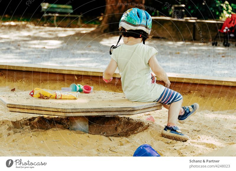 Kind mit Fahrradhelm sitzt im Sandkasten auf dem Spielplatz spielen Sandalen Kindheit Außenaufnahme Spielen Farbfoto Kindergarten Kleinkind Spielzeug