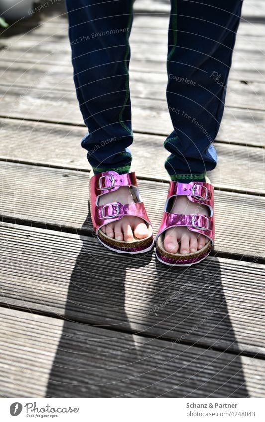 Kind mit pinken Lacksandalen Sandalen Hausschuhe Schatten Holzdielen Balkon Sommer Kindheit kindlich verlegen Zehen Füße Fuß glitzer glänzend Barfuß Beine