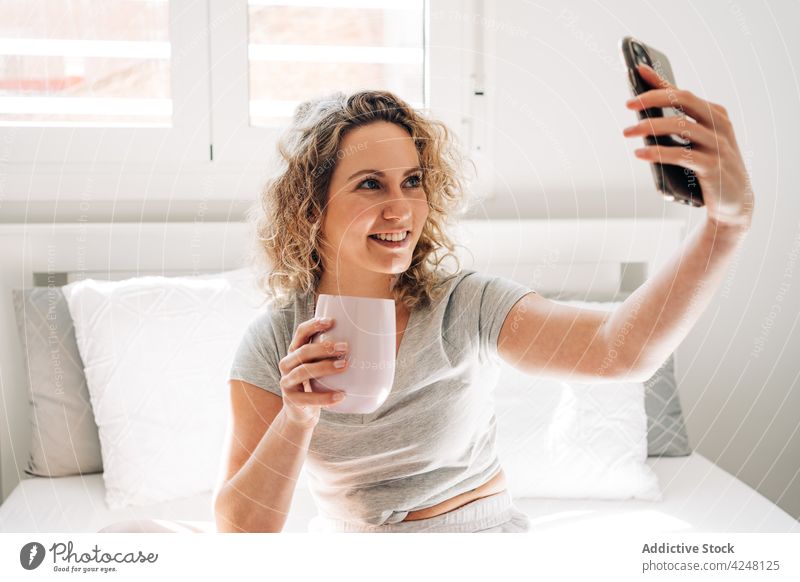 Lächelnde Frau mit Tasse nimmt Selfie auf Bett heiter Zahnfarbenes Lächeln Smartphone Becher Selbstportrait Gedächtnis Moment Schlafzimmer Gerät Mobile jung
