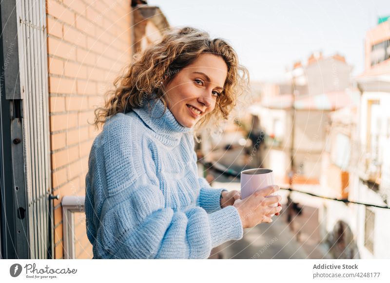 Lächelnde junge Frau trinkt Kaffee auf dem Balkon trinken Stil genießen Getränk Morgen sich[Akk] entspannen heimwärts heiß gemütlich krause Haare blond Kälte