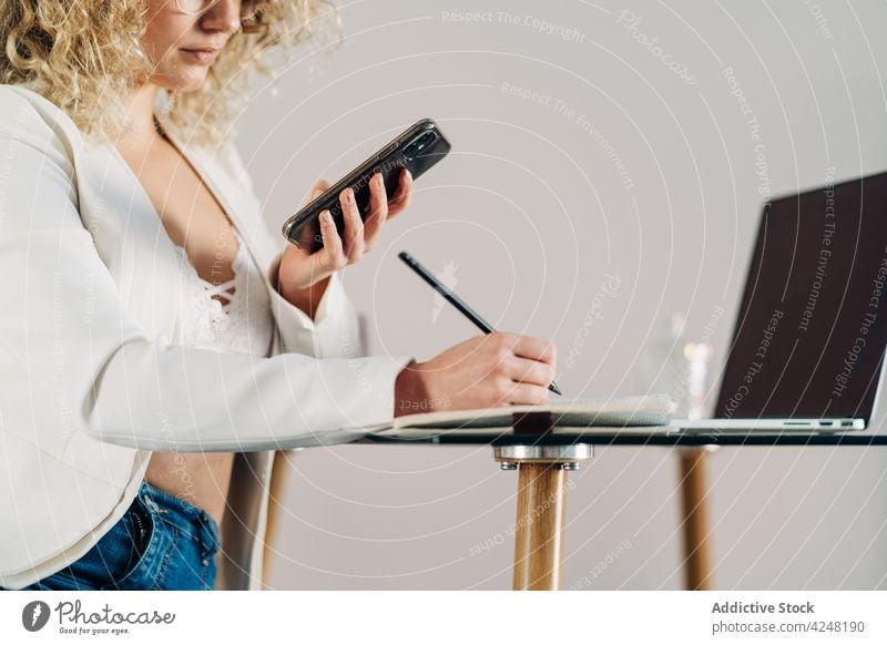 Anonyme seriöse junge Geschäftsfrau, die im Büro Notizen macht und auf ihrem Smartphone surft benutzend zur Kenntnis nehmen Arbeit Konzentration online Laptop