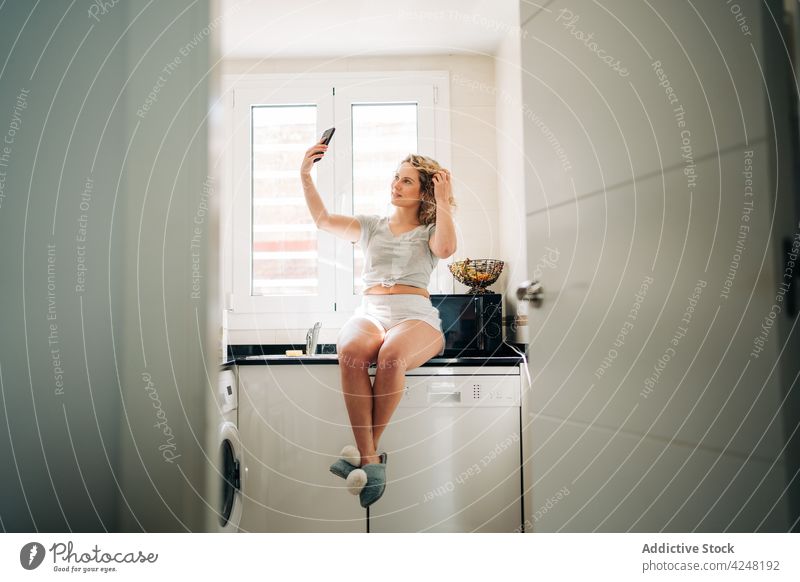 Fröhliche Frau, die ein Selfie macht und auf der Küchenzeile sitzt Haare berühren Smartphone Selbstportrait benutzend heiter fotografieren Inhalt Apparatur