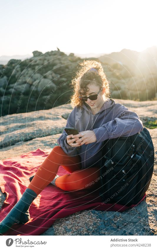 Glückliche junge Frau, die auf ihrem Smartphone Nachrichten sendet, während sie sich beim Trekking auf einem Hügel ausruht benutzend Wanderer Lächeln erkunden