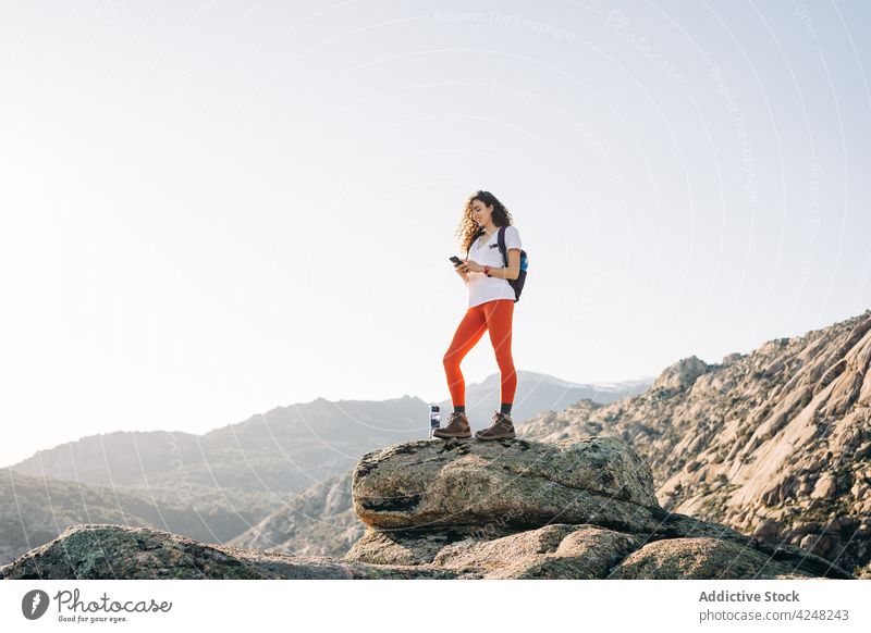 Fröhliche junge Rucksacktouristin, die auf einem Berghang im Sonnenlicht ihr Smartphone benutzt Frau Lächeln Reisender erkunden Berge u. Gebirge Trekking trendy