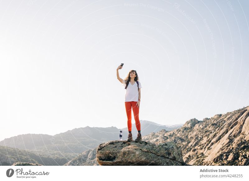 Fröhliche junge Rucksacktouristin macht ein Selfie auf einem Berghang im Sonnenlicht Frau Lächeln Reisender Smartphone erkunden Berge u. Gebirge Trekking