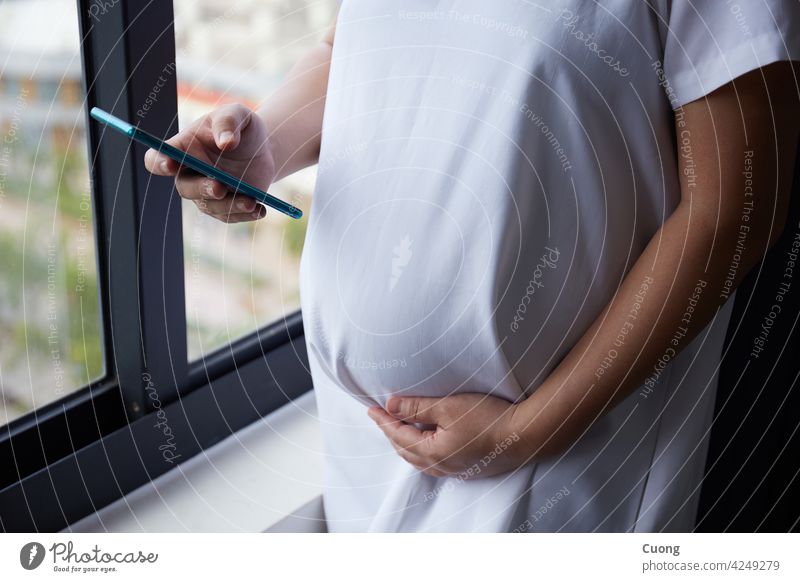 Junge asiatische schwangere Frau mit Smartphone für die Überprüfung der Nachrichten beim Stehen in der Nähe des Fensters Erwachsener App Baby schön Schlafzimmer