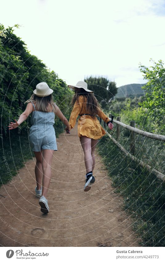 Zwei Mädchen mit Hüten, die lächelnd rückwärts einen Weg mit vielen Pflanzen entlanglaufen. Glück reden jung Lächeln Wald Frau Jugend Energie Lifestyle Park