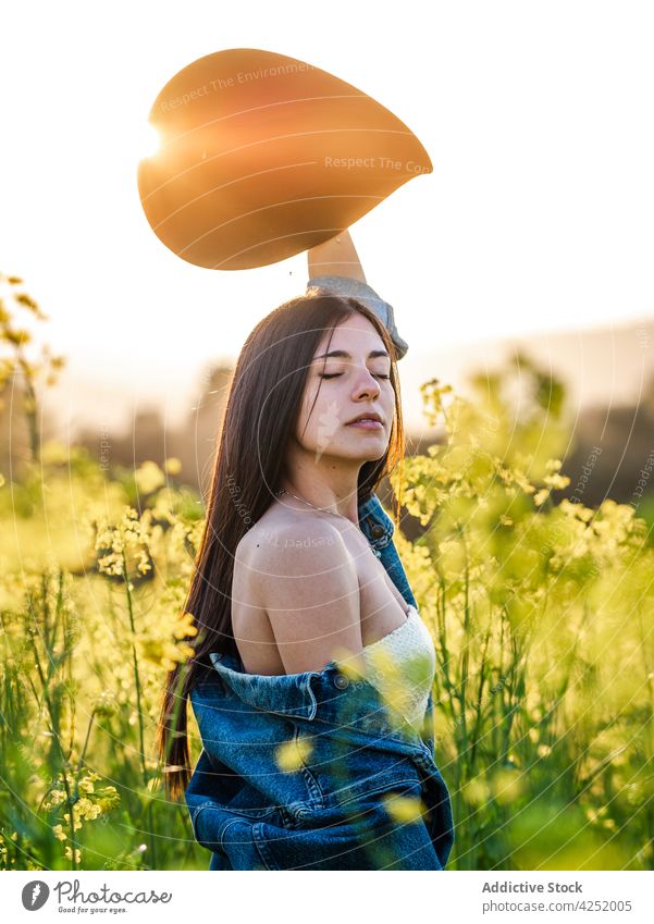 Frau hebt den Arm mit Hut und steht auf einem blühenden Feld sensibel Arme hochgezogen Augen geschlossen nackte Schultern Blüte Gelassenheit Natur Raps feminin
