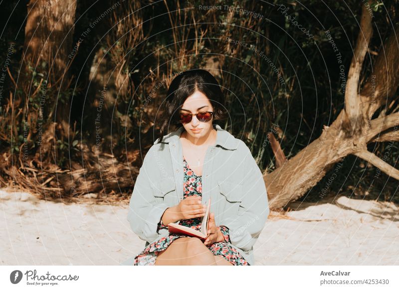Junge marokkanische Frau auf moderne Kleidung mit Sonnenbrille sitzen am Strand lesen ein Buch während eines sonnigen Tages mit Kopie Raum inspirierend und entspannen Thema mit bunten Tönen