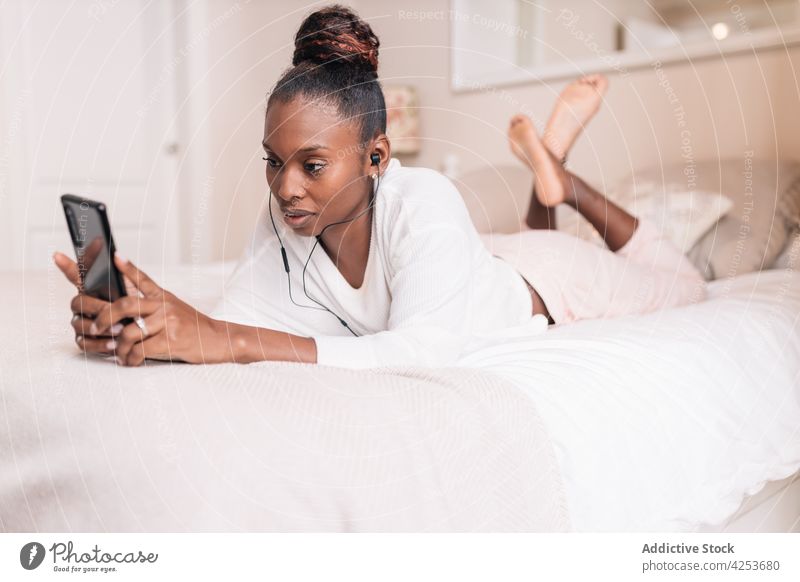 Positive junge schwarze Frau, die ein Handy benutzt und Musik hört, während sie auf dem Bett liegt Lügen Smartphone selbstsicher Lächeln zuhören
