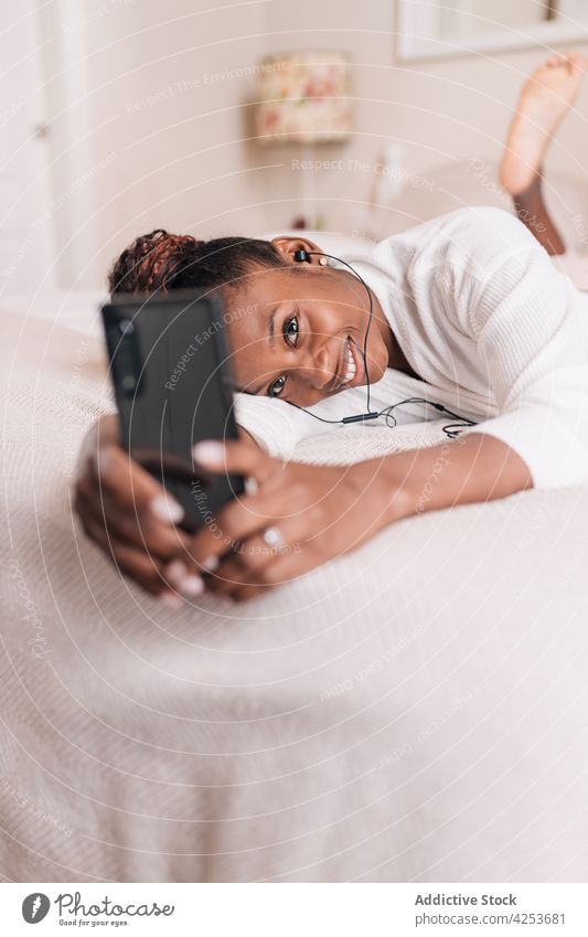 Positive junge schwarze Frau, die ein Selfie macht und Musik hört, während sie auf dem Bett liegt Lügen Smartphone selbstsicher Lächeln zuhören