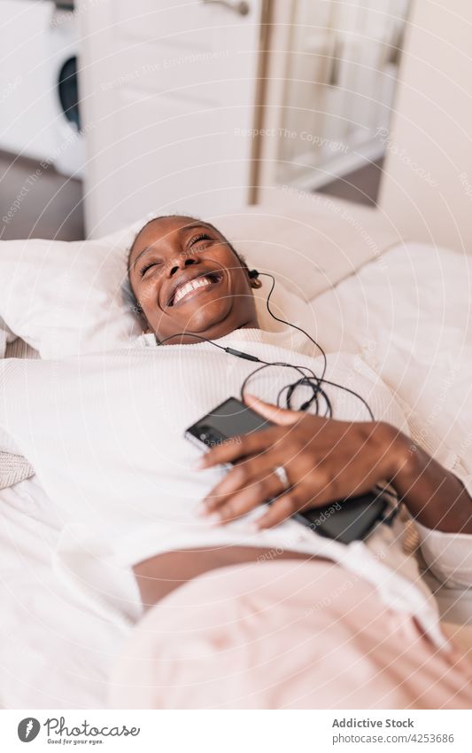 Junge ethnische Frau hört mit geschlossenen Augen Musik über Kopfhörer zuhören Augen geschlossen Lügen Bett genießen sich[Akk] entspannen Smartphone meloman