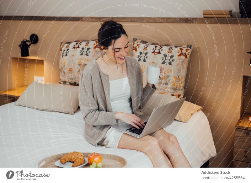 Positive Frau mit Laptop auf dem Bett Inhalt benutzend Morgen Frühstück Schlafzimmer Browsen heimisch Gerät Internet sitzen online Apparatur Lächeln Netbook