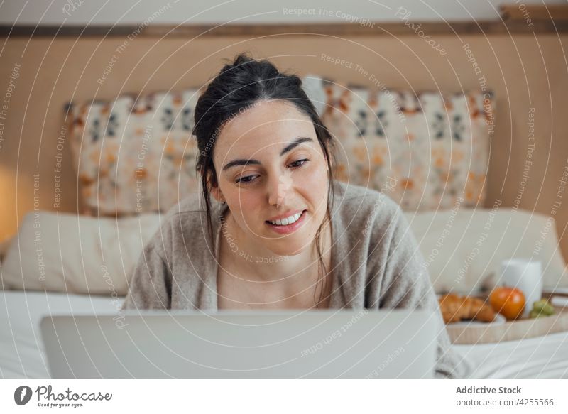 Positive Frau mit Laptop auf dem Bett Inhalt benutzend Morgen Frühstück Schlafzimmer Browsen Lügen heimisch Gerät Internet online Apparatur Lächeln Netbook