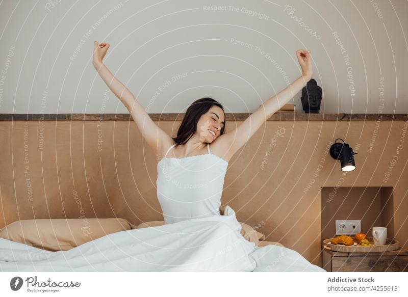 Lächelnde Frau streckt die Arme nach gutem Schlaf auf dem Bett aus aufwachen Dehnung Augen geschlossen Zahnfarbenes Lächeln Inhalt Schlafzimmer Morgen froh