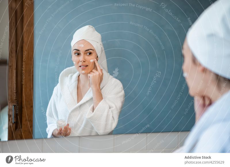Frau im Bademantel beim Eincremen des Gesichts im Badezimmer Sahne bewerben froh Hautpflege Spiegel Gesichtsbehandlung Routine verjüngen Feuchtigkeit