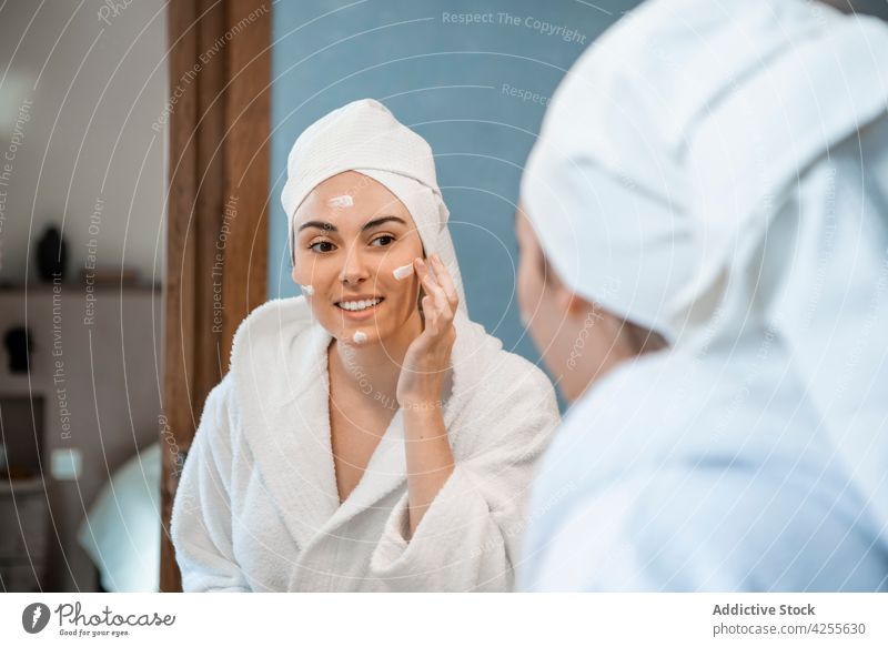 Frau im Bademantel beim Eincremen des Gesichts im Badezimmer Sahne bewerben froh Hautpflege Spiegel Gesichtsbehandlung Routine verjüngen Feuchtigkeit