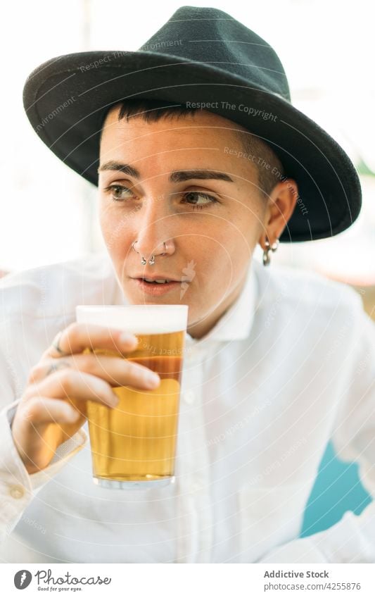 Lächelnde queere Frau trinkt Bier am Tisch Wildfang Schnaps Geschmack Alkohol Glas brauen Transgender agender Identität schäumen Getränk Becher Erfrischung