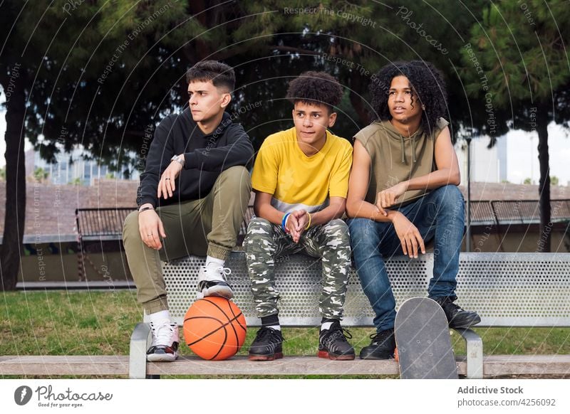 Multiethnische Jugendliche sitzen auf einer Bank im Park Männer Freund achtsam Interesse Fokus Basketball Zusammensein Zeitvertreib Skateboard männlich