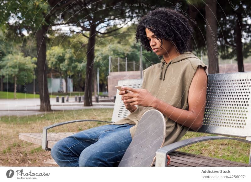 Schwarzer Mann sitzt auf einer Bank und benutzt ein Smartphone benutzend Internet Skateboard Pause Nachricht Park Texten Anschluss männlich Browsen Surfen