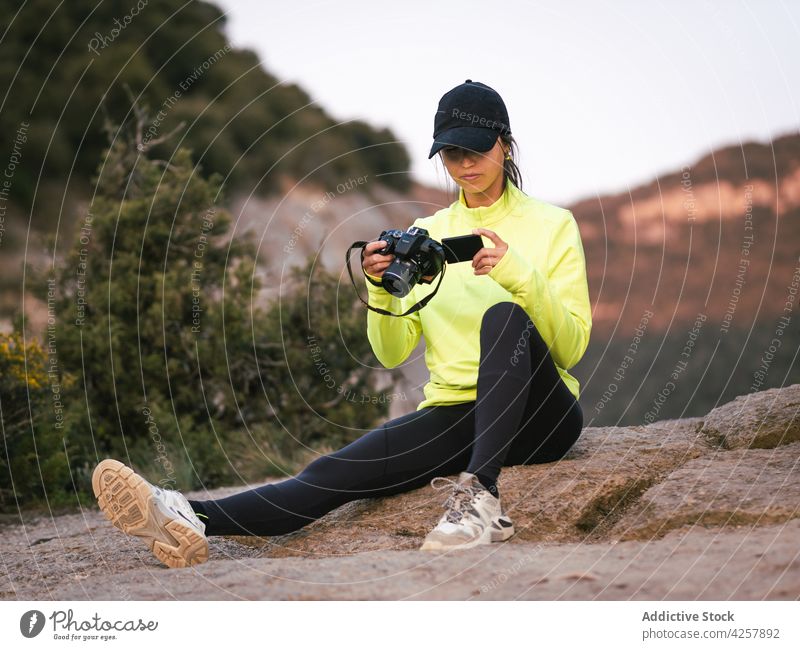 Junge ethnische Frau, die während einer Wanderung auf einem Stein ausruht und sich ein Video ansieht zuschauen Reisender Klippe Natur Trekking Tal Camcorder