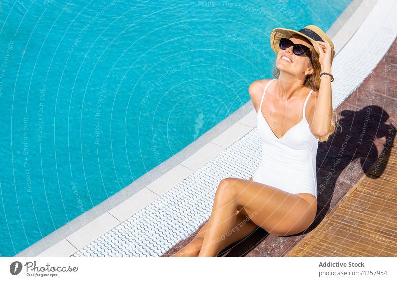 Glückliche Frau am Pool sitzend und ein Sonnenbad nehmend Badeanzug Beckenrand Hut Resort Kälte Urlaub Sonnenbrille Badebekleidung ruhen Paradies Bräune