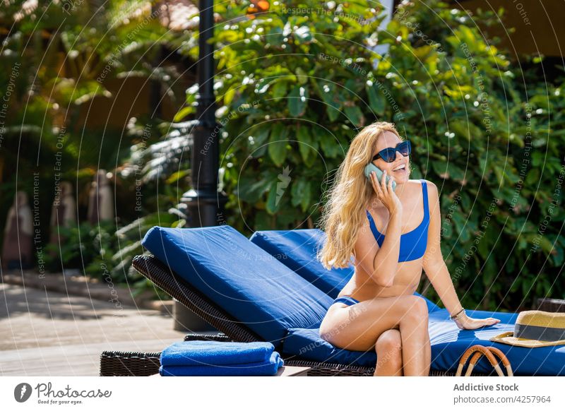 Fröhliche Frau im Bikini, die auf einem Liegestuhl am Pool mit ihrem Smartphone spricht Telefonanruf Sonnenbad Beckenrand freudig Zahnfarbenes Lächeln reden