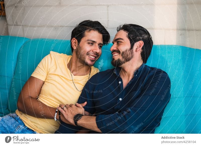 Lächelndes ethnisches Paar von Schwulen, die sich auf der Couch an den Händen halten schwul Händchenhalten Partnerschaft Liebe Inhalt räkeln Zeit verbringen