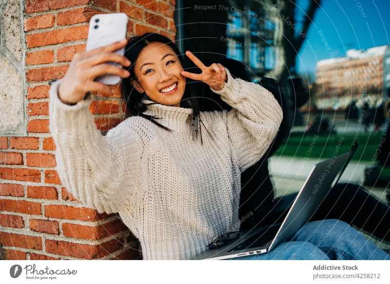 Positive asiatische Millennials machen ein Selfie mit ihrem Laptop auf der Straße Frau Lächeln Smartphone Arbeit freiberuflich heiter zwei Finger Internet