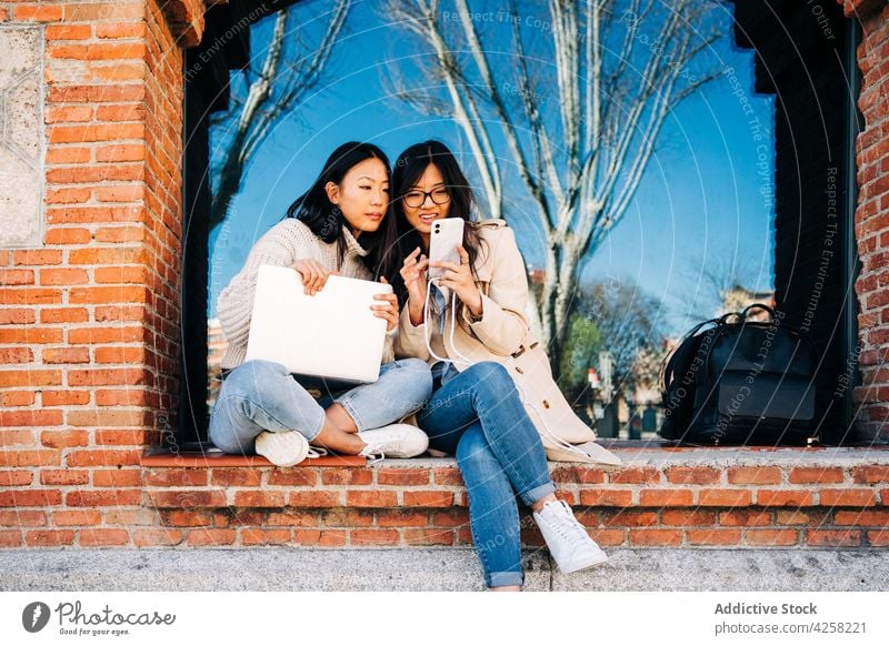 Positiv eingestellte junge ethnische Frauen, die am Laptop in der Stadt telefonieren Freude Zeit verbringen bester Freund Stil Zusammensein expressiv Browsen