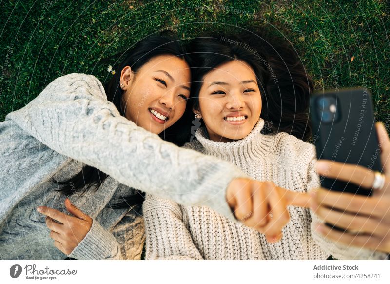 Joyful jungen ethnischen weiblichen Freunde liegen auf Rasen und zeigt Zungen, während der Aufnahme selfie Frauen Selfie Zunge zeigen Lügen Freude Glück