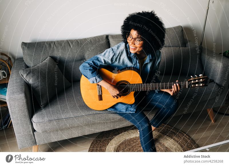 Positive kreative schwarze Frau spielt Gitarre auf dem Sofa im Wohnzimmer positiv Lächeln heiter akustisch heimwärts spielen Musik Brille lässig Instrument