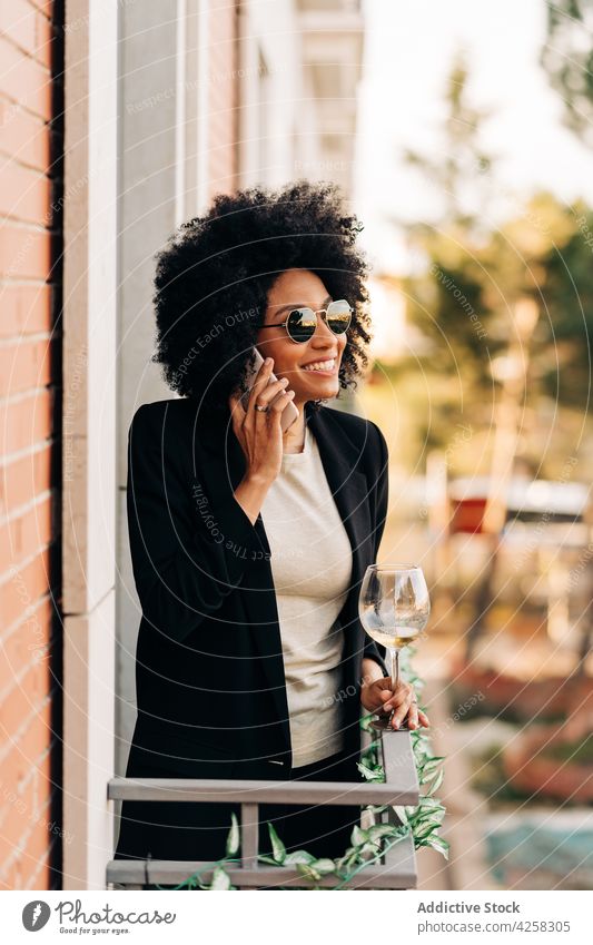 Stilvolle schwarze Frau mit trendiger Sonnenbrille im Gespräch mit Smartphone Anzug elegant reden Glas Telefonanruf Wein positiv Lächeln heiter Mobile Alkohol