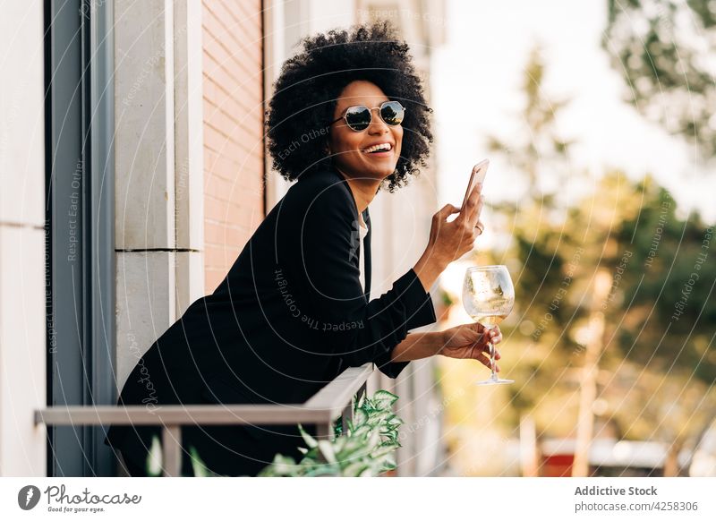 Stilvolle schwarze Frau mit trendiger Sonnenbrille und Smartphone Anzug elegant Glas Wein positiv Lächeln heiter Mobile Telefon Alkohol Balkon Dame