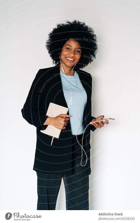 Fröhliche schwarze Frau mit Notizbuch und Smartphone hört Musik heiter Lächeln Glück Kopfhörer Notebook Mobile Telefon positiv elegant Anzug formal Outfit