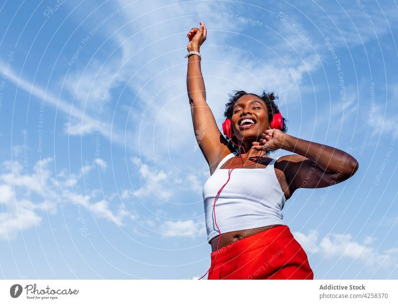 Aufgeregte schwarze Frau, die mit Kopfhörern Musik hört und tanzt Tanzen zuhören Freude Spaß haben Blauer Himmel Melodie genießen Arme hochgezogen Glück jung