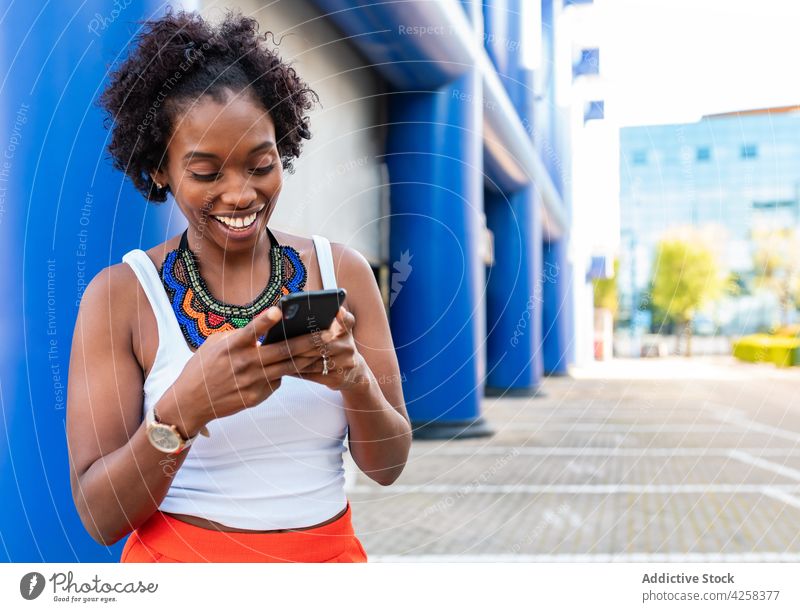 Fröhliche schwarze Frau, die ein Smartphone benutzt und auf der Straße geht benutzend freudig Großstadt heiter schlendern Browsen Gerät Training schlank Energie