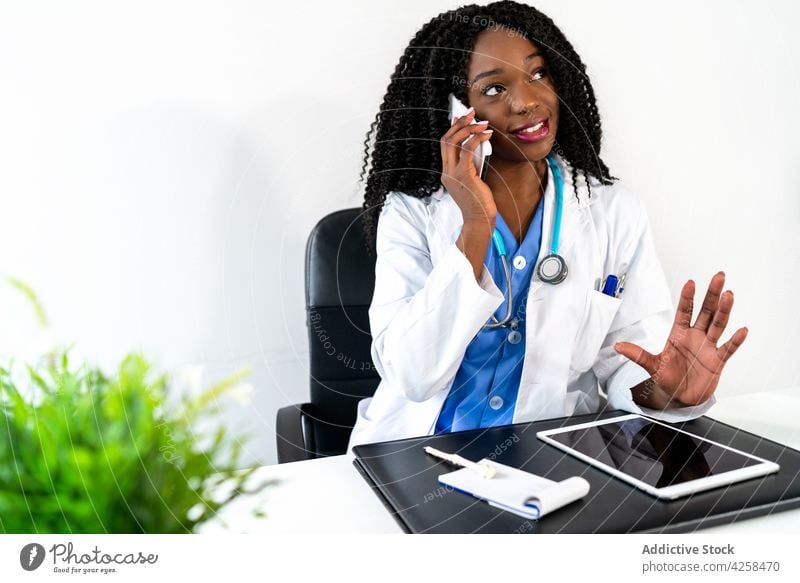 Lächelnde schwarze Frau, die im Büro über ihr Smartphone spricht Arzt reden benutzend konsultieren Klinik Telefonanruf Gesundheitswesen Uniform Stethoskop