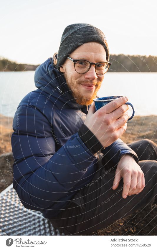 Glücklicher junger Mann trinkt ein heißes Getränk beim Camping am Seeufer trinken Kaffee Ufer Lager Wanderer reisen ruhen positiv Lächeln männlich Vollbart Stil