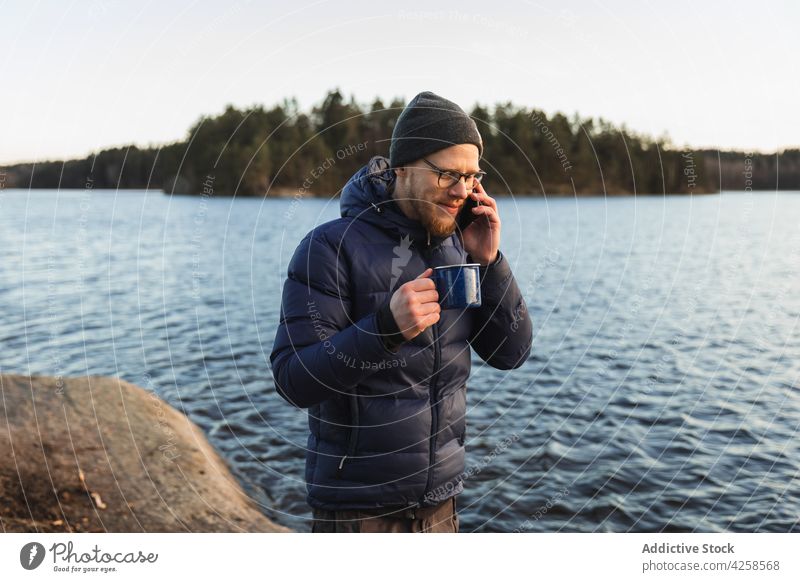 Junger männlicher Reisender führt ein Telefongespräch und trinkt Kaffee am Seeufer Mann trinken Telefonanruf Getränk Smartphone Wanderer Wald Natur