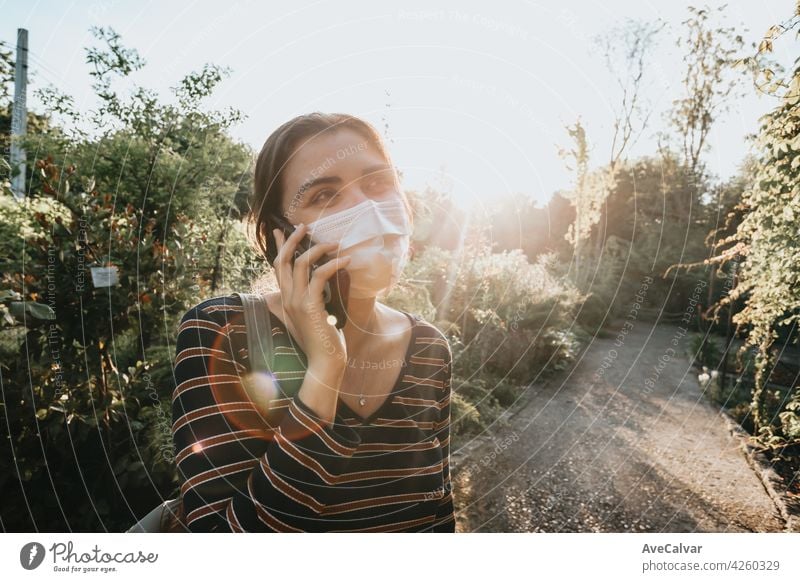 Junge Frau mit einer Gesichtsmaske beim Telefonieren in einem Garten an einem super sonnigen Tag Textfreiraum Lächeln soziale Netzwerke Kaukasier genießen