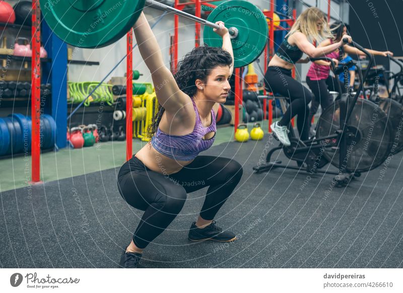 Frau übt Gewichtheben in der Turnhalle Kurzhantel Greifen Arme hoch Tatkraft Überwindung Überkopfhocke Sportlerin selbstbewusst Zug Stärke Training