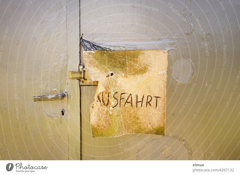 Ein quadratisches, eingerissenes Stück Leder mit der gekritzelten Aufschrift " AUSFAHRT " verdeckt den Türriegel an einer geschlossenen, khakifarbenen Garagentür aus Metall, deren Rostflecken überstrichen wurden / Provisorium