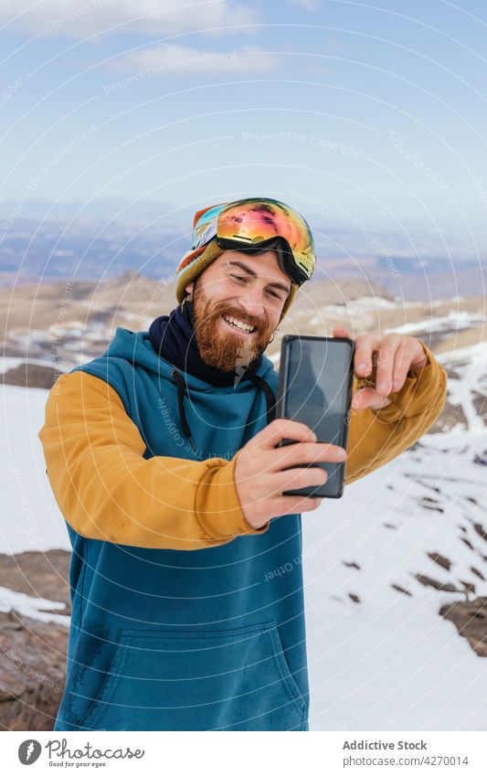 Lächelnder Sportler, der in den winterlichen Bergen ein Selfie mit seinem Smartphone macht heiter Gedächtnis Moment Berge u. Gebirge Schnee Hipster benutzend