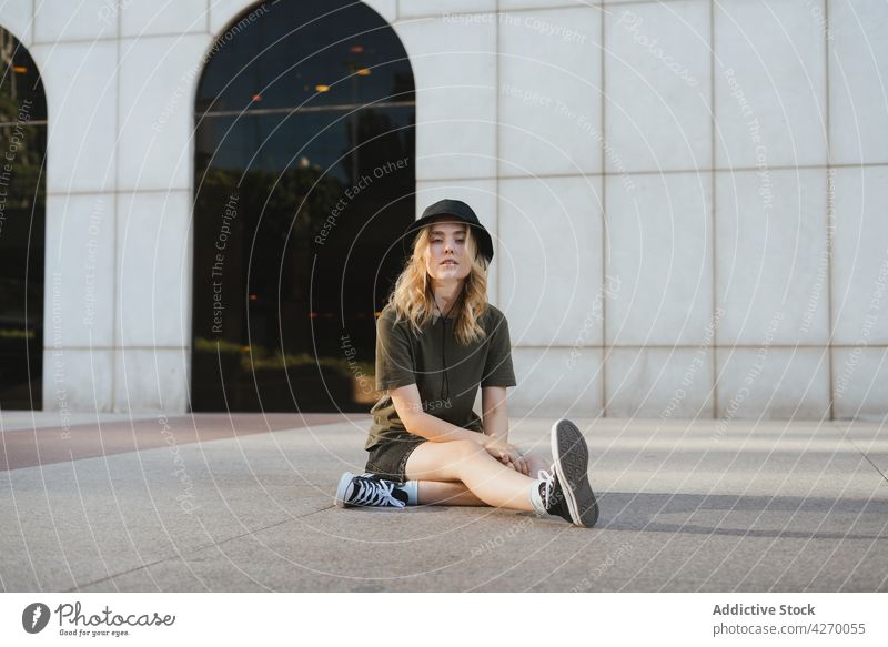 Coole Frau mit Hut, die sich auf dem Bürgersteig der Stadt ausruht cool Individualität Streetstyle Beine gekreuzt Zeitgenosse Generation Laufsteg Porträt