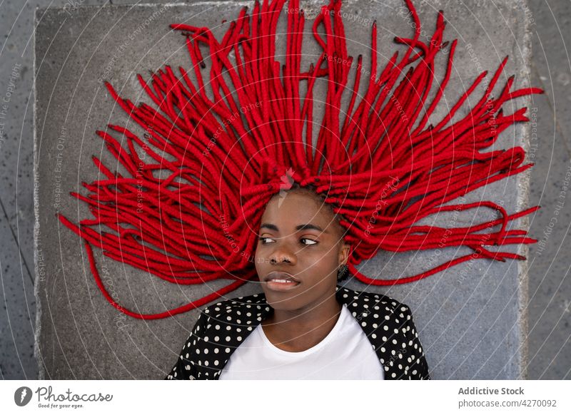 Schwarze Frau mit heller Frisur liegt auf einer Betonfläche Geflecht rot cool sorgenfrei Stil ernst Starrer Blick Lügen Individualität Persönlichkeit lebhaft