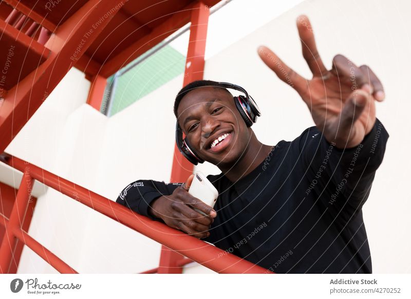 Fröhlicher schwarzer Mann mit Kopfhörern und V-Zeichen v-Zeichen Zahnfarbenes Lächeln heiter zwei Finger zeigen zuhören gestikulieren sorgenfrei zufrieden