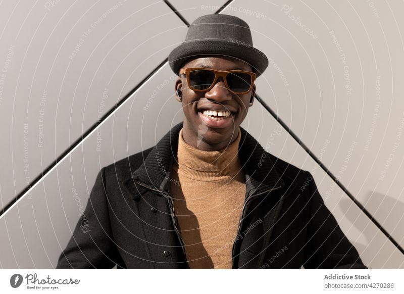 Stilvoller fröhlicher schwarzer Mann vor weißer Wand stehend Outfit selbstbewusst Vorschein trendy cool gut gekleidet Hut Sonnenbrille Individualität