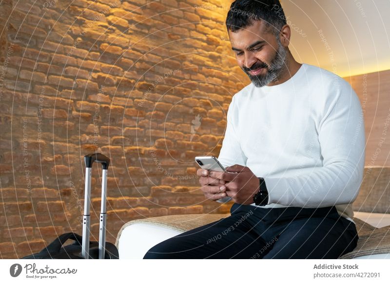 Lächelnder ethnischer Mann sitzt auf dem Bett und schreibt eine SMS auf seinem Smartphone Tourist benutzend Hotel Koffer Ausflug Urlaub positiv männlich reisen